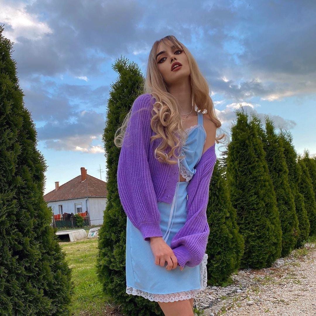Model wearing HAIRCUBE blonde synthetic wig with bangs styled in loose curls with purple cardigan and blue dress