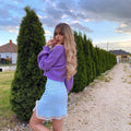 Blonde synthetic wig with bangs worn by a woman in a purple sweater and light blue skirt outdoors
